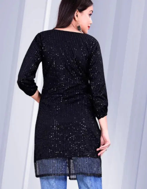 Woman wearing a black sequined dress with blue jeans against a white curtain background