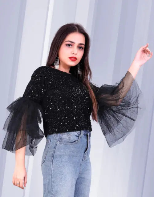 Woman wearing a black sequin top with sheer sleeves against a light curtain background