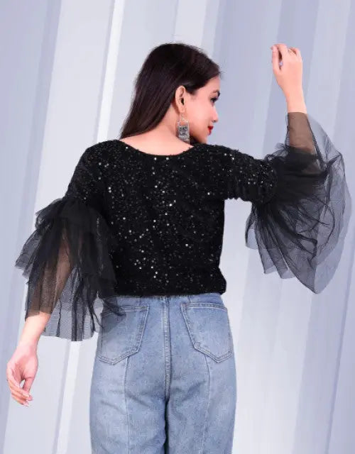 Woman wearing a black sequin top with sheer sleeves against a light gray curtain.